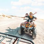 Štvorkolka jazdiaca cez rozľahlú púšť s píšťalovými dunami na pozadí